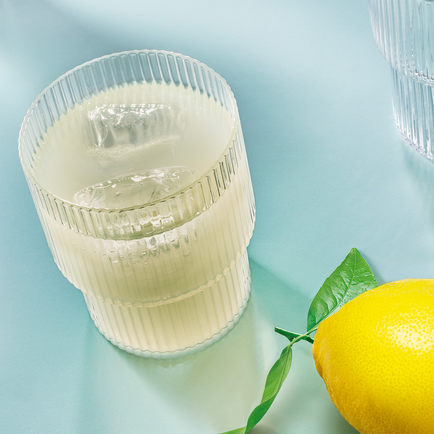 Ein Glas frischer Limonade der Adelholzener Primella Limonaden mit einer Zitrone auf blauem Untergrund
