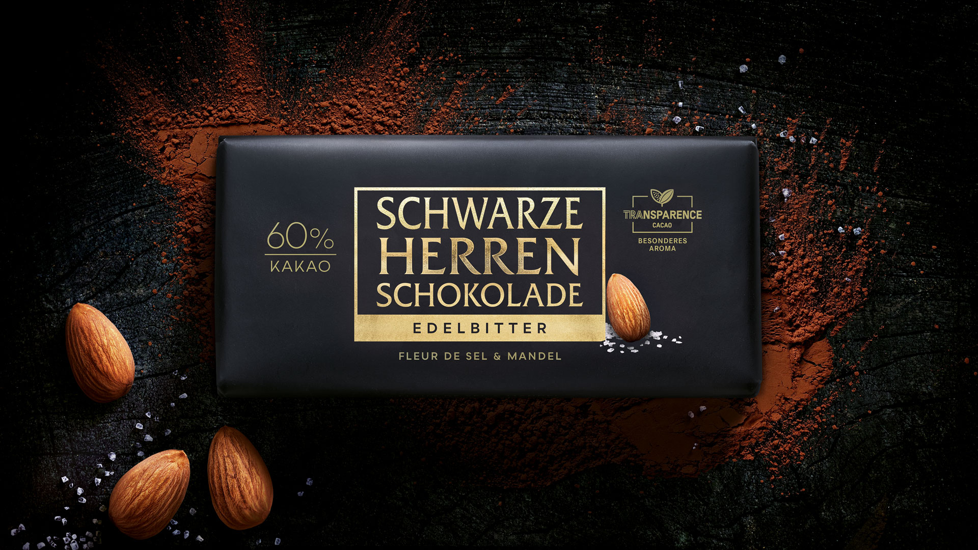 Edelbitterschokolade „Schwarze Herren“ mit 60 % Kakao, Fleur de Sel und Mandeln auf dunklem Hintergrund mit Kakaopulver.