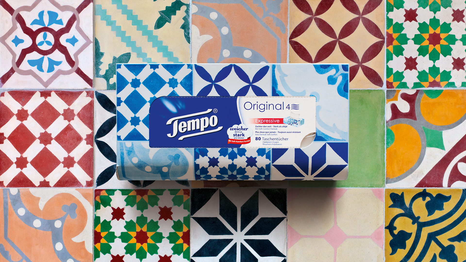 Designstarke Inszenierung der Tempo Original Taschentuchbox vor einem bunten Mosaikfliesen-Hintergrund: Die Box mit blauen Ornamenten und prominenter Markenkennzeichnung liegt im Zentrum des Bildes – visuell harmonisch eingebettet in ein Patchwork aus geometrisch gemusterten Fliesen.