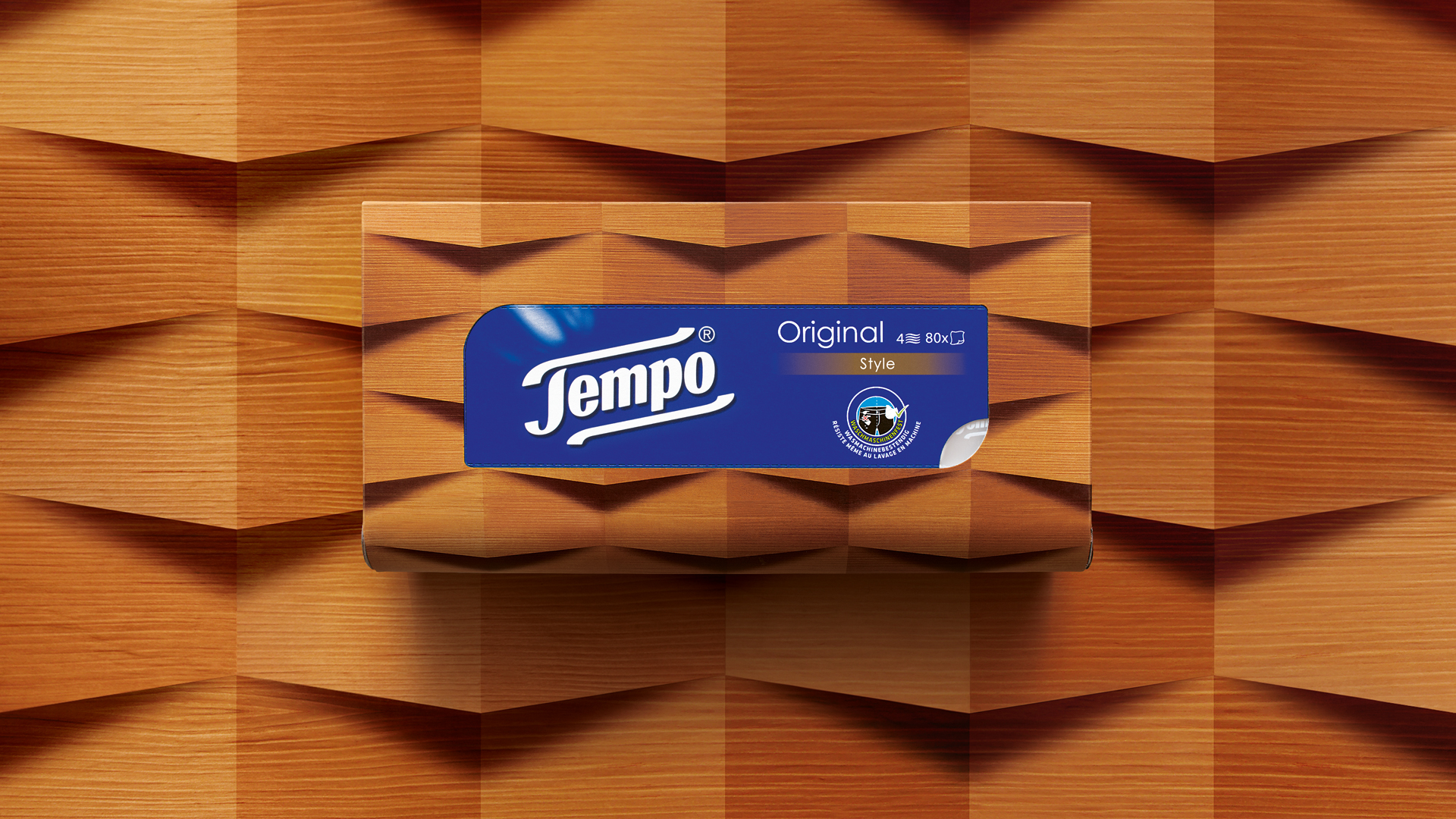 Tempo Original Taschentuchbox in der Variante ‚Style‘ vor einem geometrisch gefalteten Holzdekor – die Verpackung fügt sich nahtlos in das warme, skulpturale Hintergrundmuster aus 3D-Holzoptik ein und unterstreicht den hochwertigen, designorientierten Look.