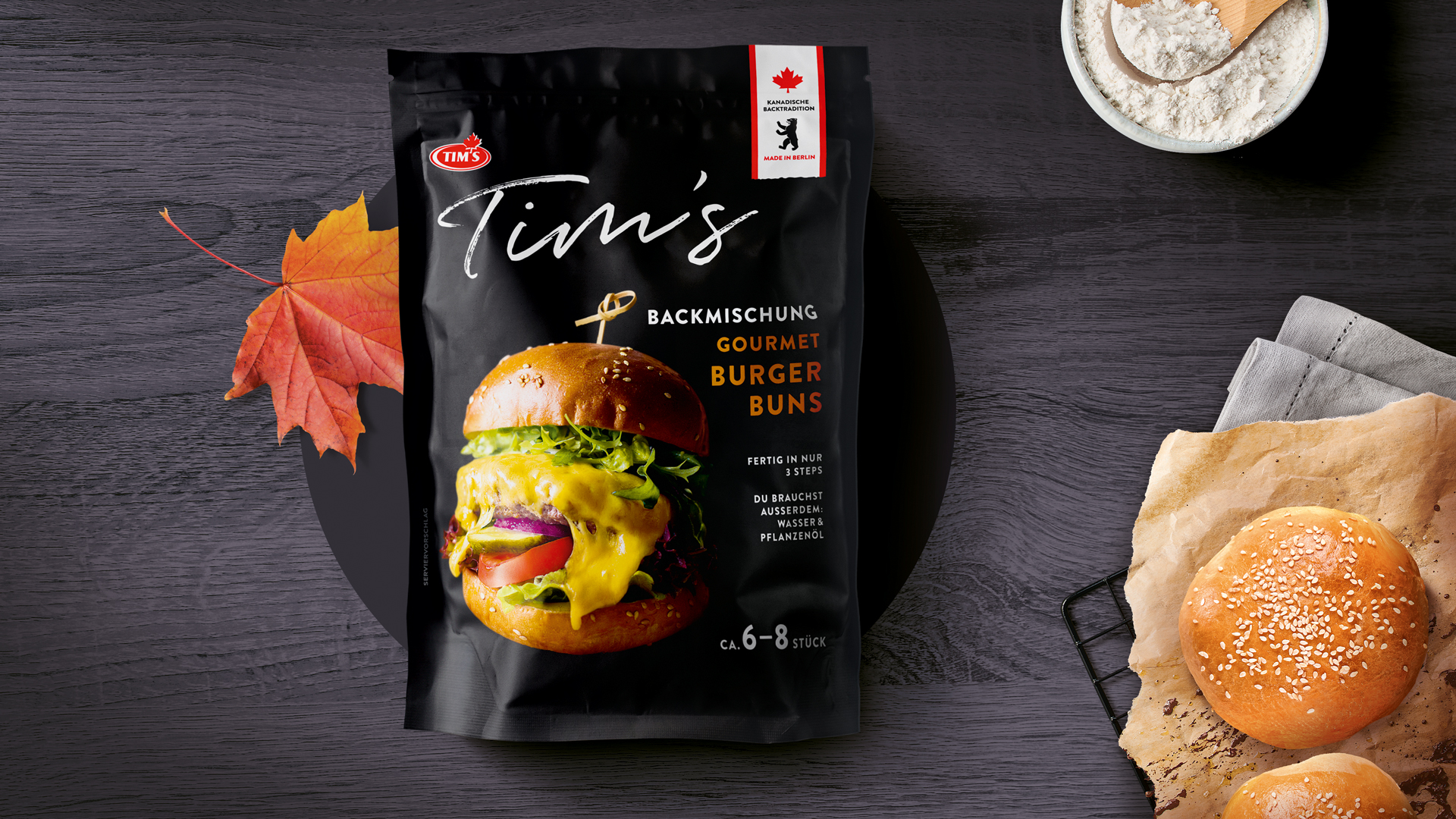 Stilvoll inszeniertes Verpackungsdesign für Tim’s Gourmet Burger Buns: Schwarze Tüte mit appetitlichem Burgermotiv, Ahornblatt als kanadischer Akzent und frisch gebackene Buns auf dunklem Holzuntergrund.